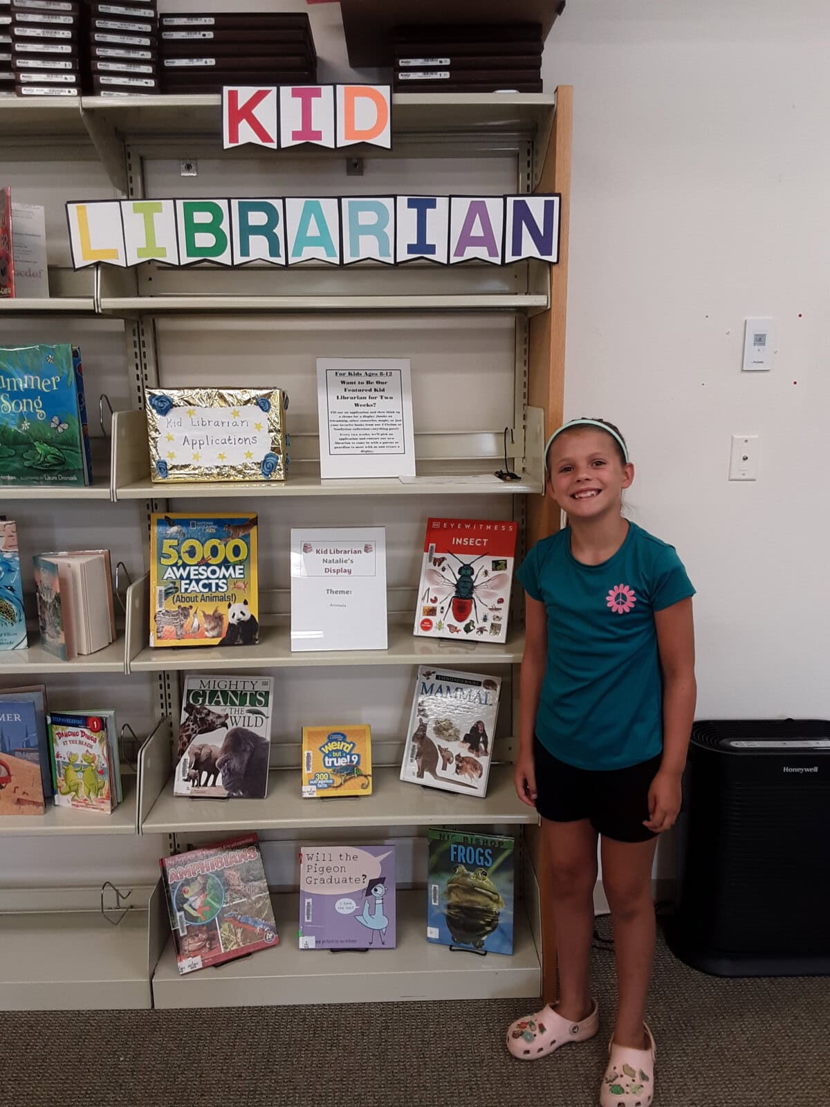 A young girl stands smiling by a bookshelf labeled “Kid Librarian,” featuring childrens books and a display sign. She wears a teal shirt, shorts, and a headband in a library setting.