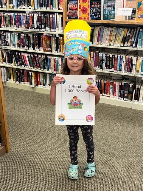 A young girl wearing a colorful 1,000 Books Before Kindergarten hat and glasses stands in a library, proudly holding a sign that says I Read 1,000 Books. Bookshelves filled with books are in the background.