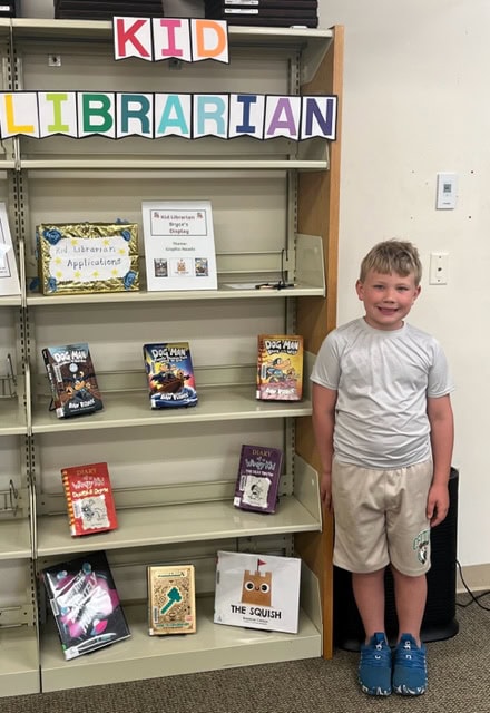 A young boy stands smiling beside a library display labeled Kid Librarian, featuring colorful books and a sign for Kid Librarian applications on mostly empty shelves.