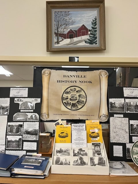 Display of historical materials titled Danville History Nook, featuring old photos, pamphlets, and maps. Above, a framed painting shows a red barn and snowy landscape. Pamphlets and binders are on the table.