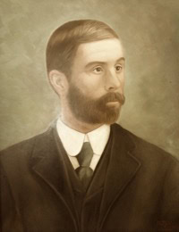 A sepia-toned portrait of a man with short brown hair, a full beard, and mustache, wearing a dark suit, white shirt, and tie, set against a soft, neutral background.