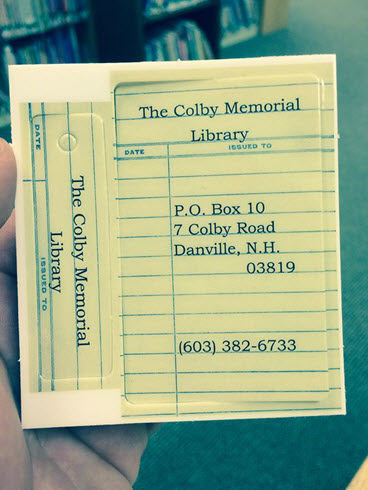A hand holds a library card pocket and card from The Colby Memorial Library in Danville, NH, displaying the library’s address and phone number, styled to look like an old checkout card. Bookshelves are blurred in the background.